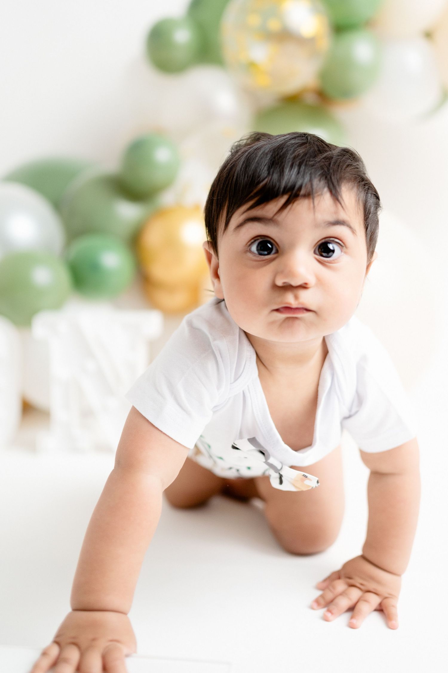 Baby crawling on white floor with green and gold balloon arch decoration.