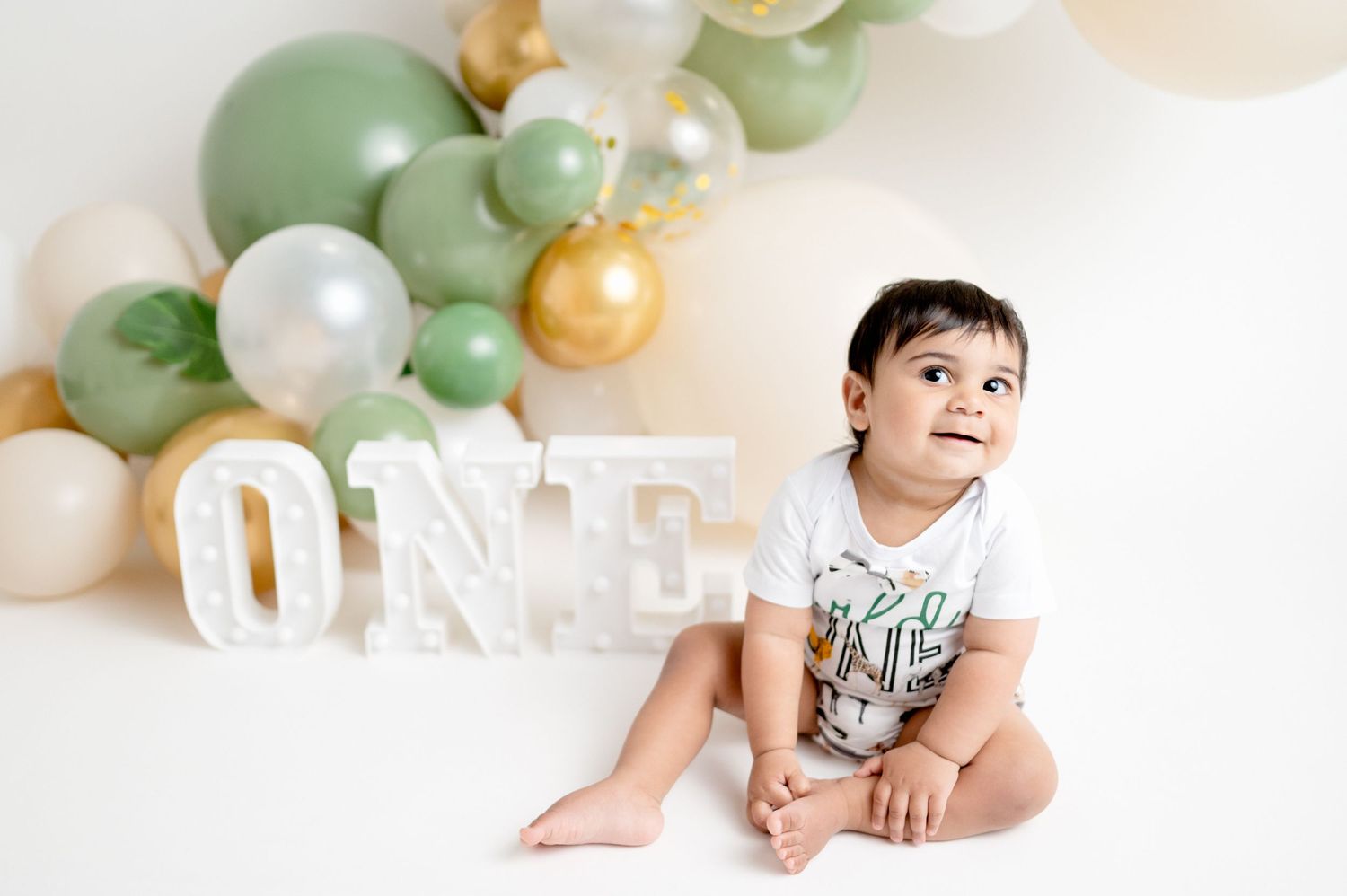 Baby in white onesie sitting beside ONE letters with elegant balloon garland backdrop.
