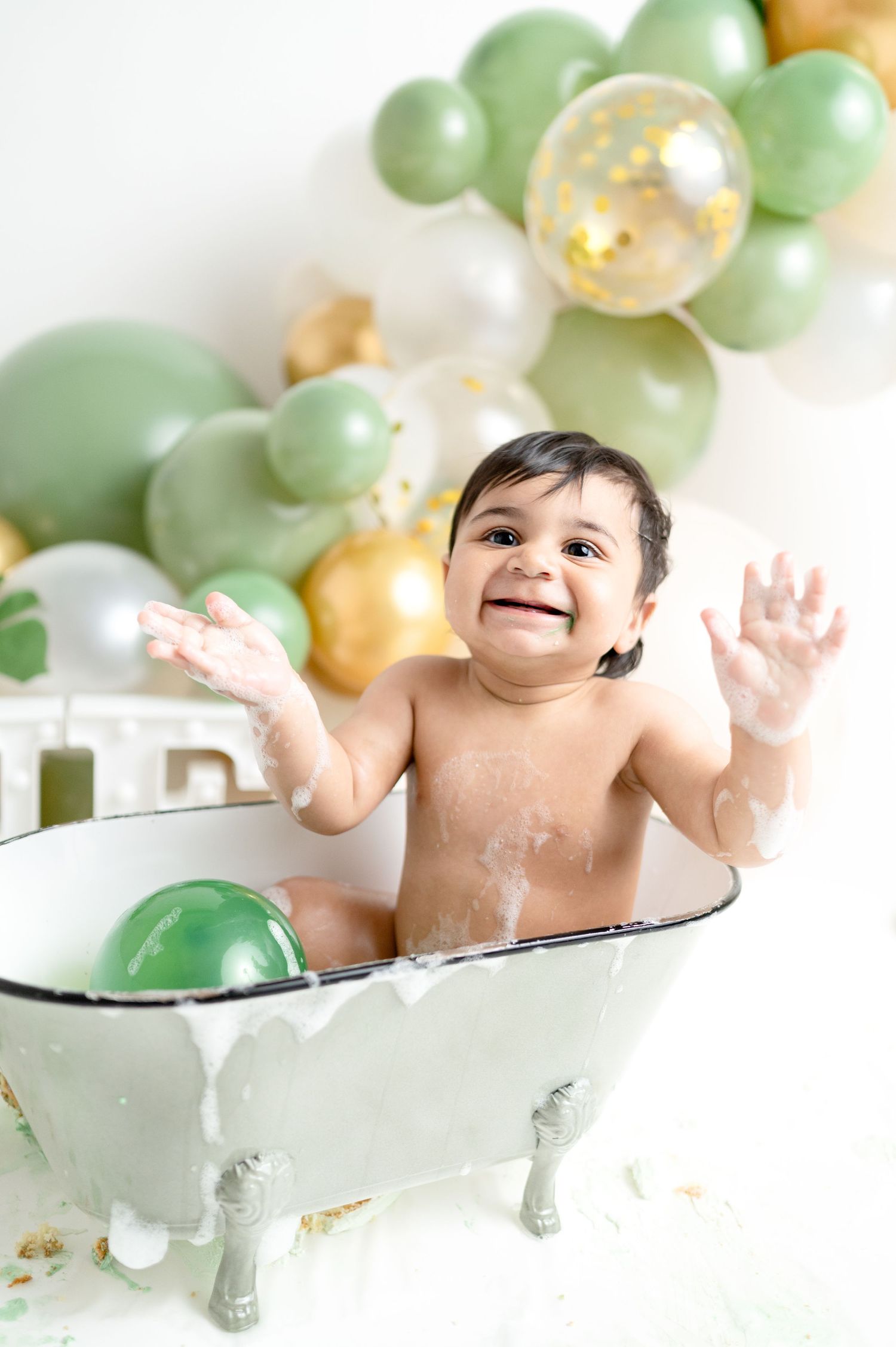 Joyful baby playing in vintage bathtub with soap and festive balloon decorations.