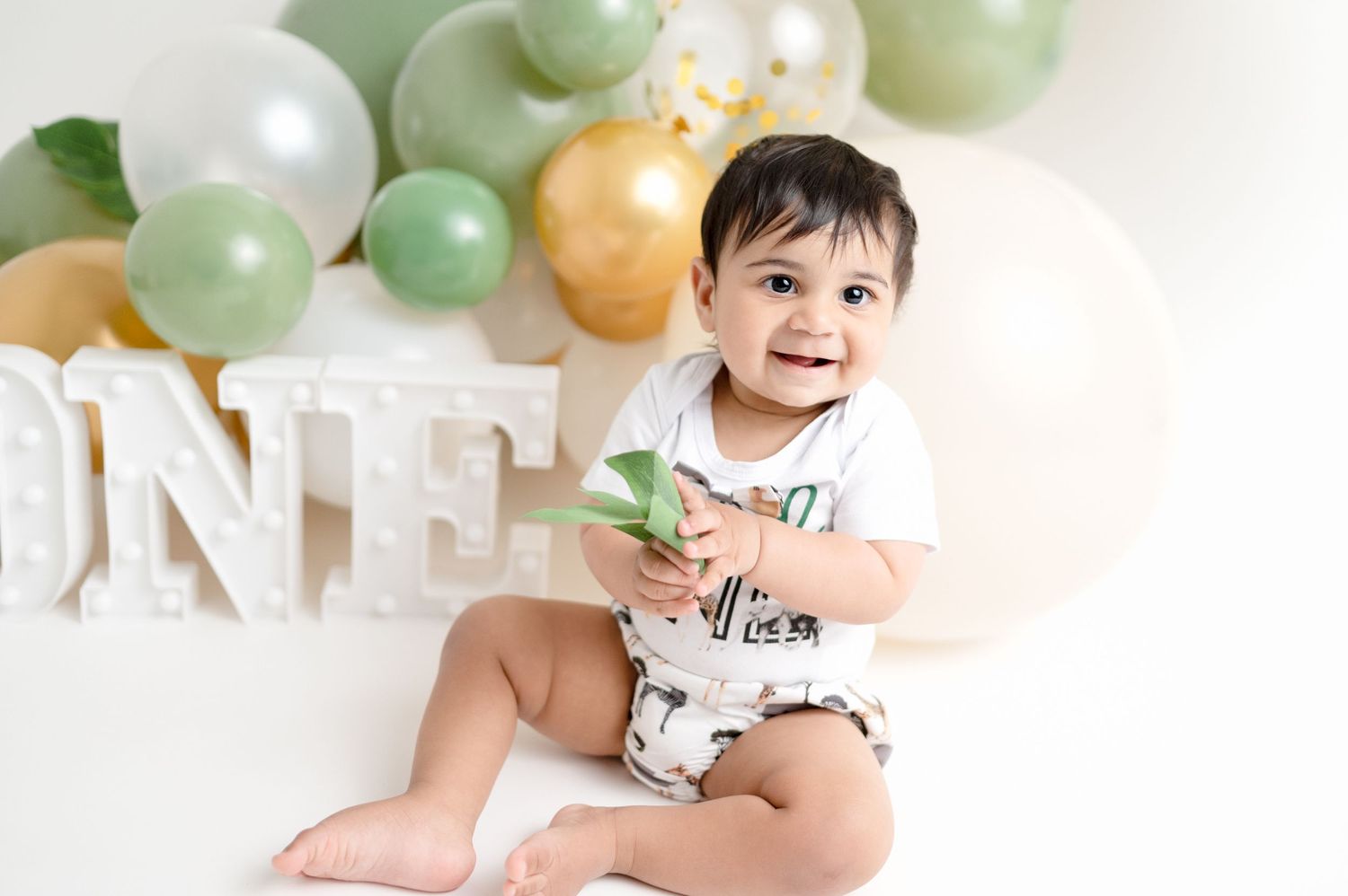 A first birthday photoshoot setup with white letters and sage green and gold balloon decorations.