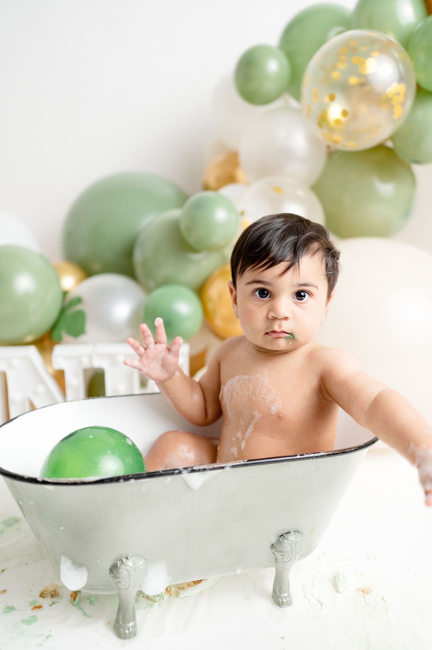 Playful baby splashing in antique bathtub with green and gold balloons.