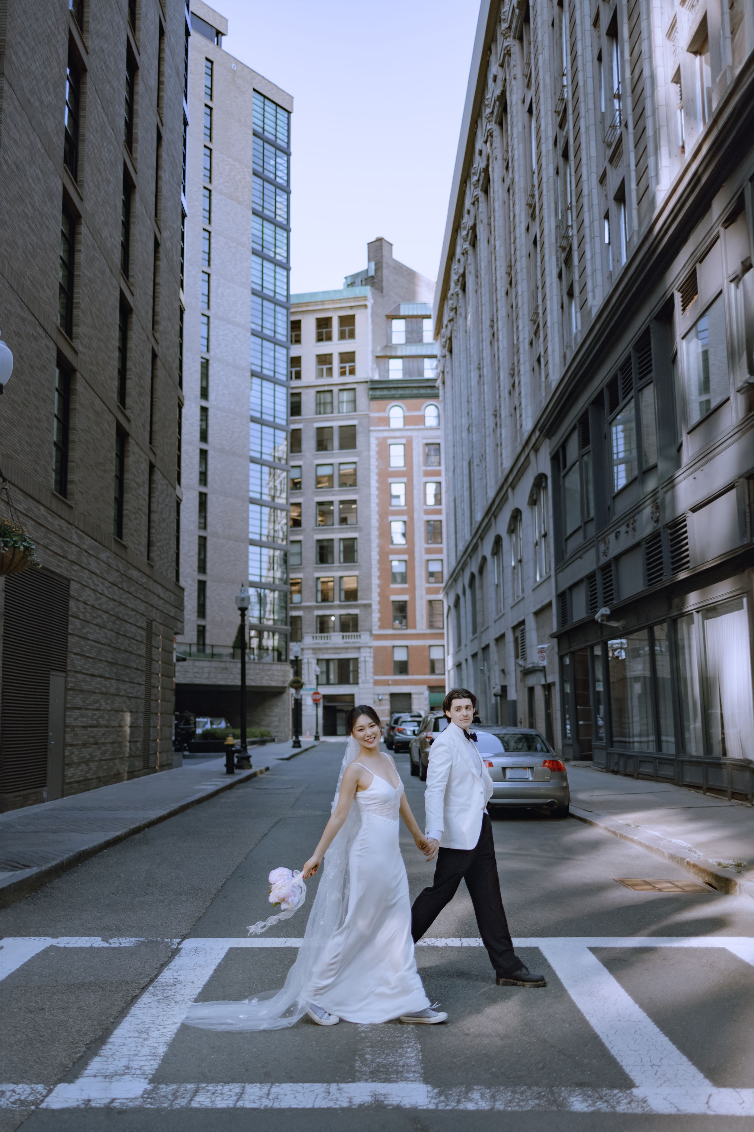 A playful wedding photo of a couple crossing a city street between tall buildings.