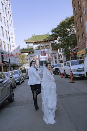 A couple in wedding attire walking on a city street with traditional Chinese architecture in the background.