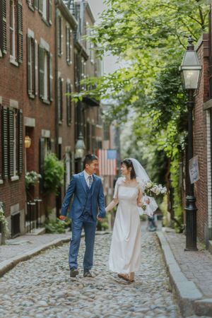 A wedding couple walking down a charming cobblestone street in Boston with brick buildings and greenery.