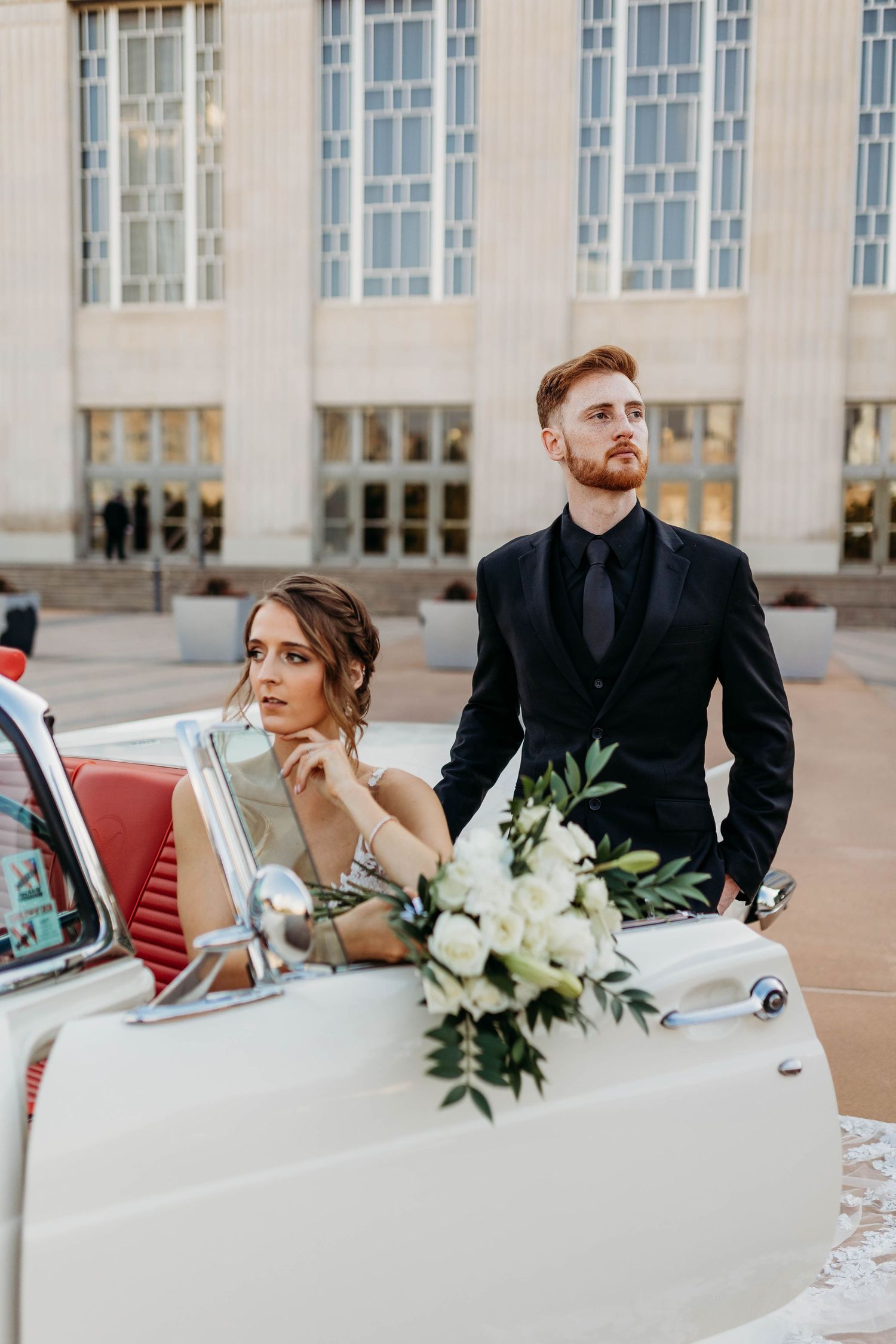 Wedding couple posing with vintage white car and floral arrangements.