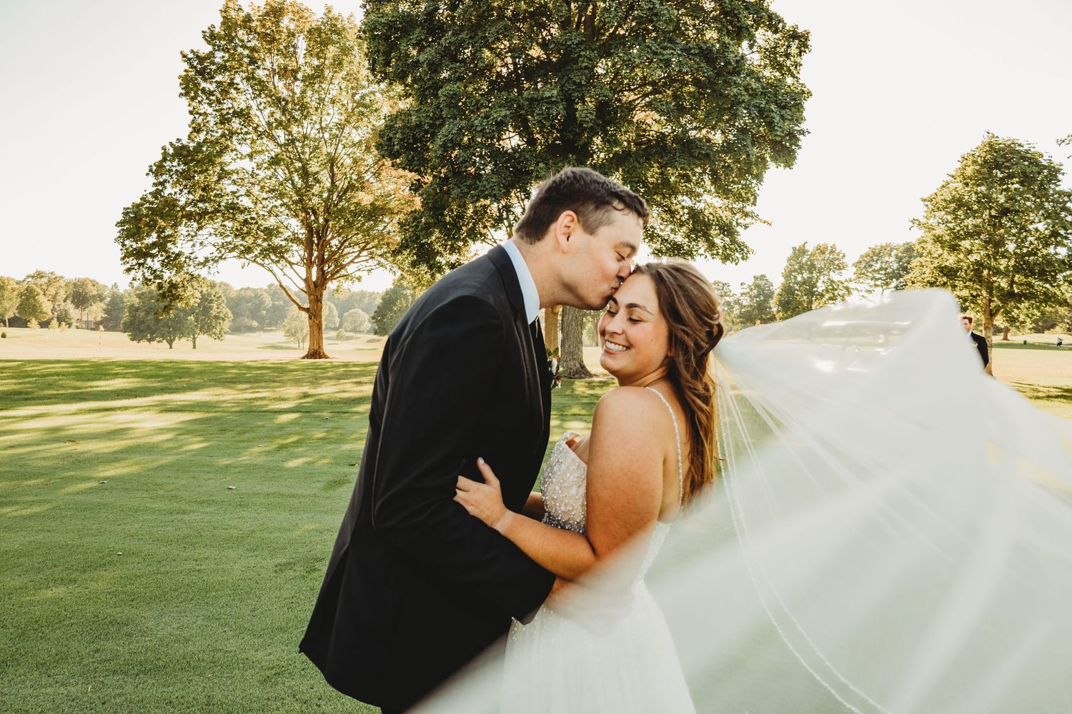 Romantic wedding couple embraces on a sunny golf course with flowing veil and natural lighting.