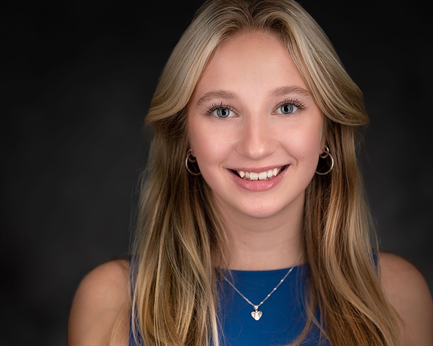 Professional portrait of young woman in blue top with necklace against dark background.