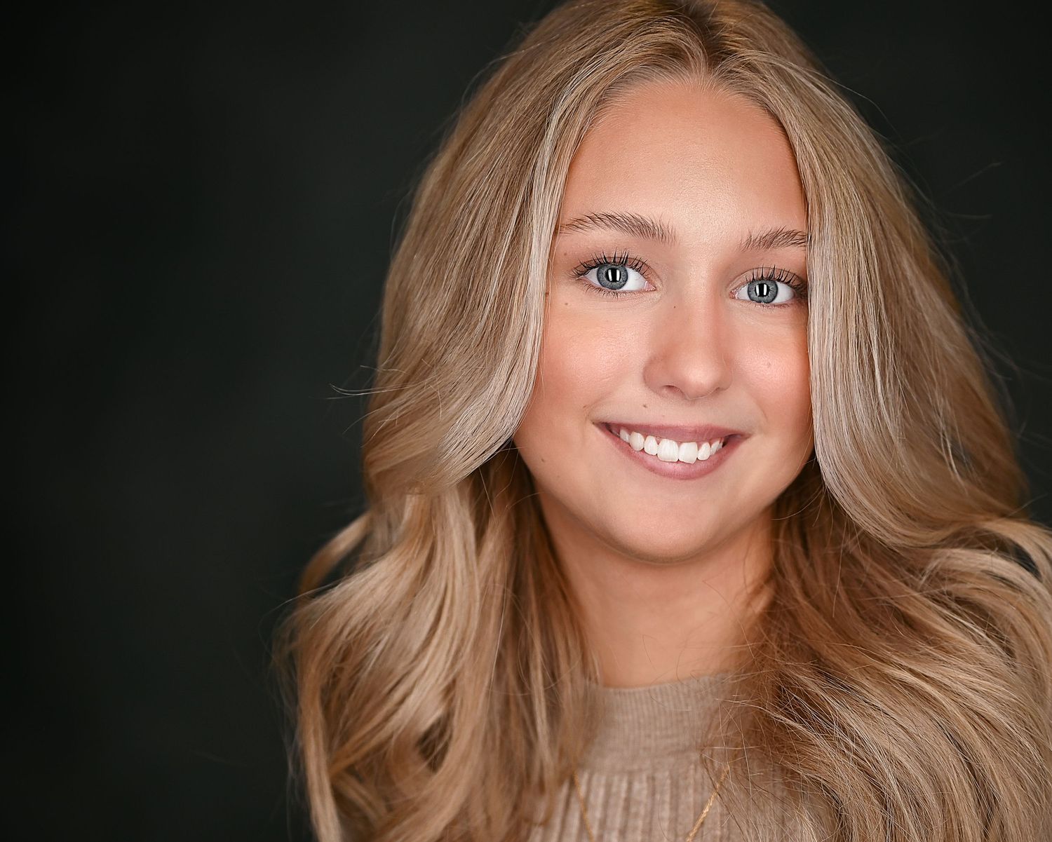 Beauty headshot of blonde woman in beige sweater with warm smile against dark backdrop.