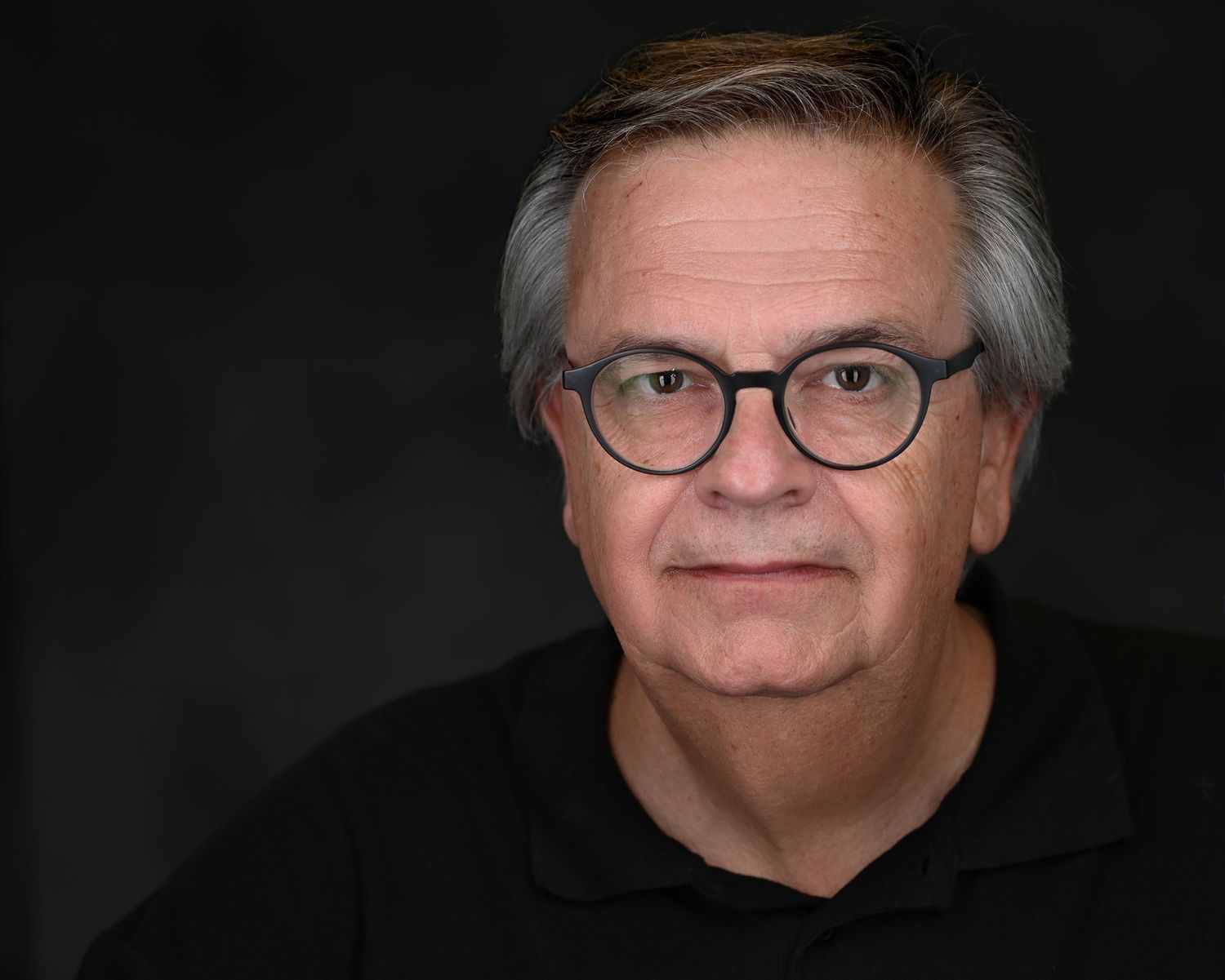 Professional headshot of mature person in black shirt and glasses against dark background.