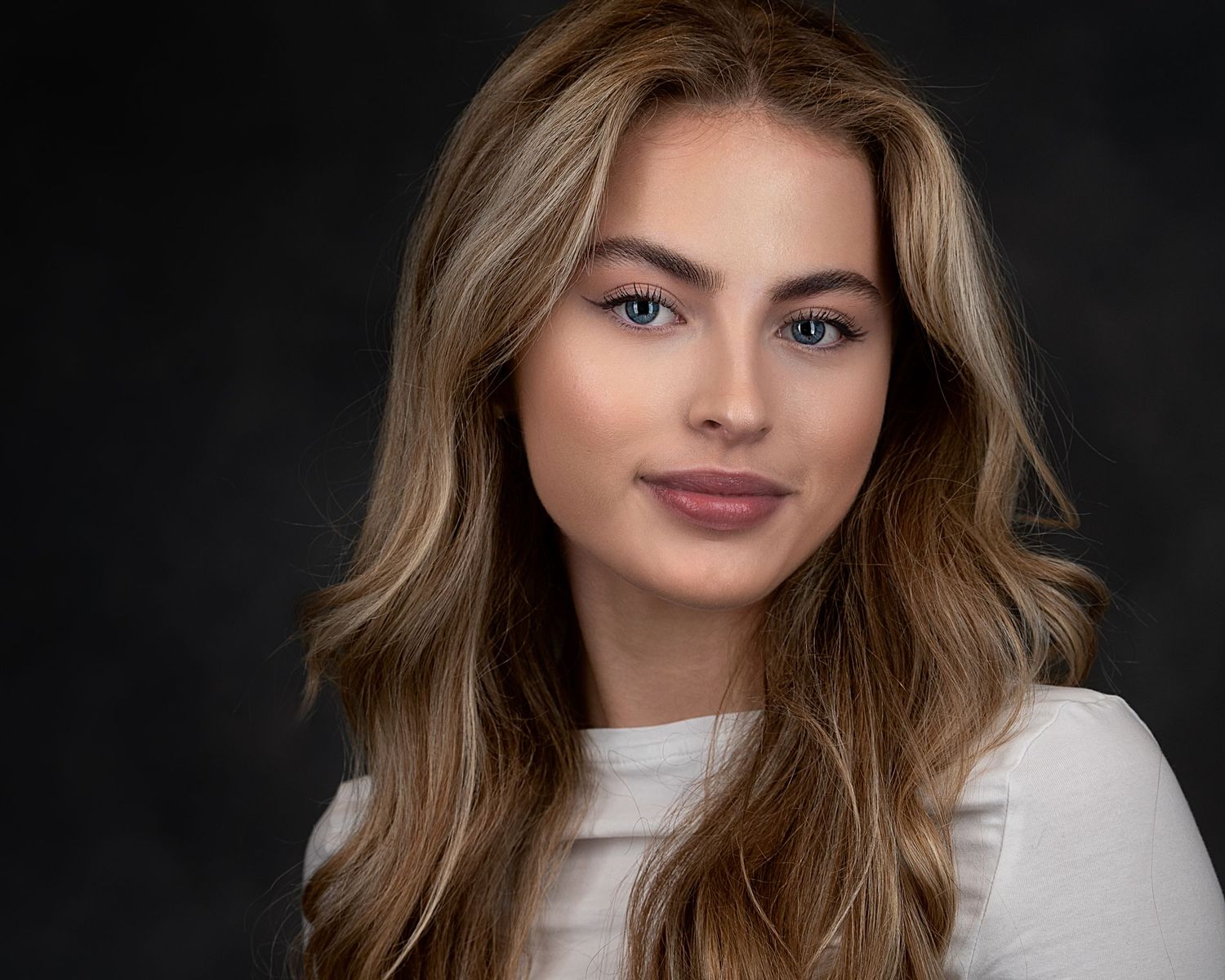 Professional beauty portrait of woman with flowing hair in white top against dark background.