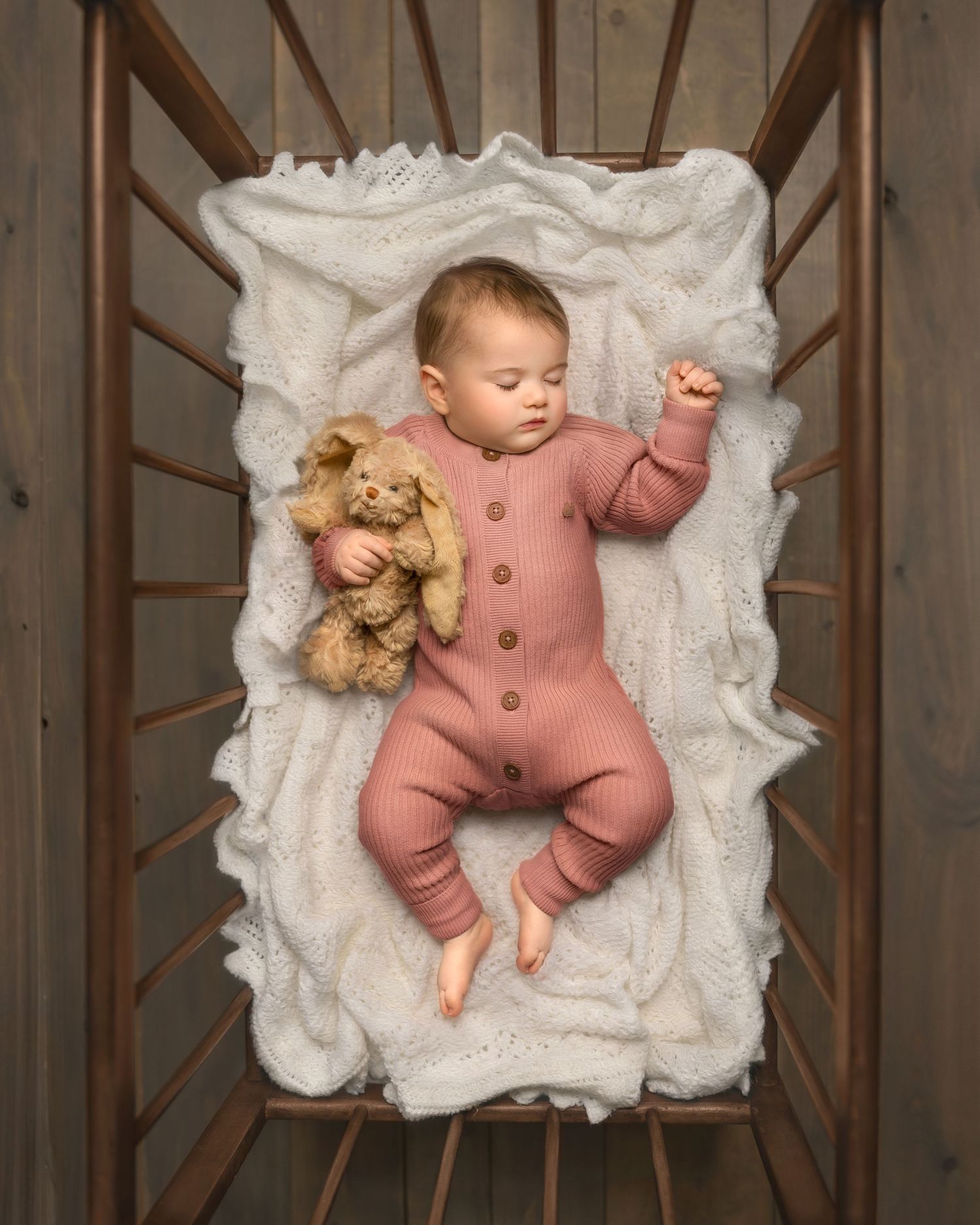 Overhead view of a sleeping baby in pink romper lying in wooden crib with soft white blanket and teddy bear.