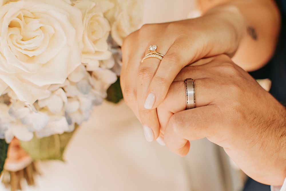 Close-up details of wedding rings and hands with white roses in the background.