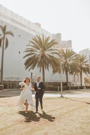 Newlyweds walking and laughing together on a sunny path lined with palm trees.