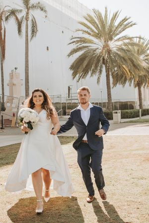 Playful wedding couple runs along palm tree-lined path in bright sunlight.