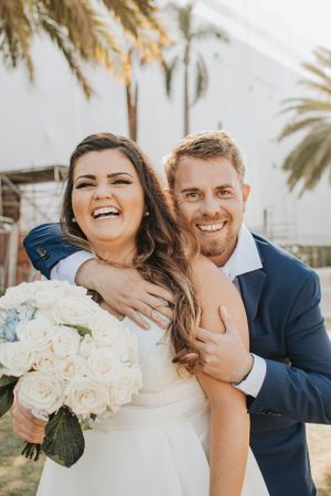 Joyful wedding couple embraces outdoors with white floral bouquet among palm trees.