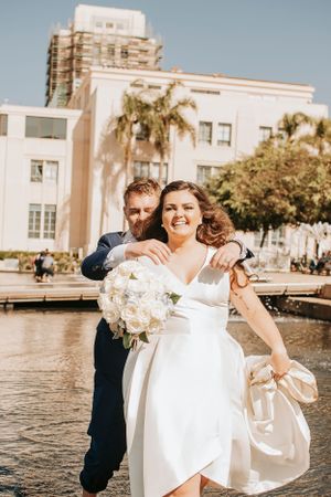 A romantic wedding couple poses by a fountain with palm trees and white buildings in the background.