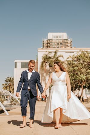 A wedding couple walks barefoot together on a sunny day with urban architecture behind them.