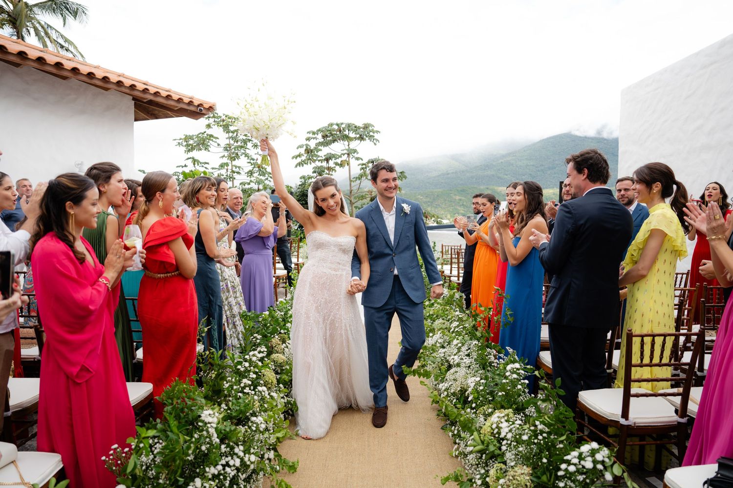 Newlyweds walk down aisle lined with colorfully dressed guests at outdoor garden ceremony.