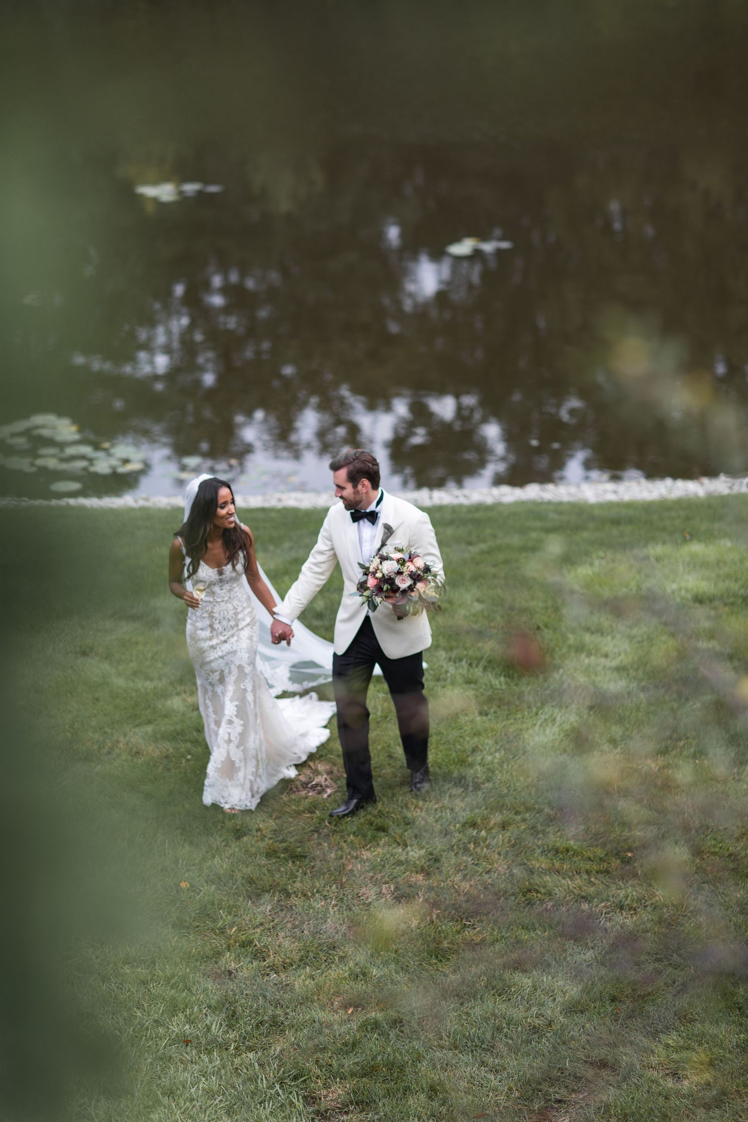 A whimsical wedding photo of a couple walking through a grassy field near water.