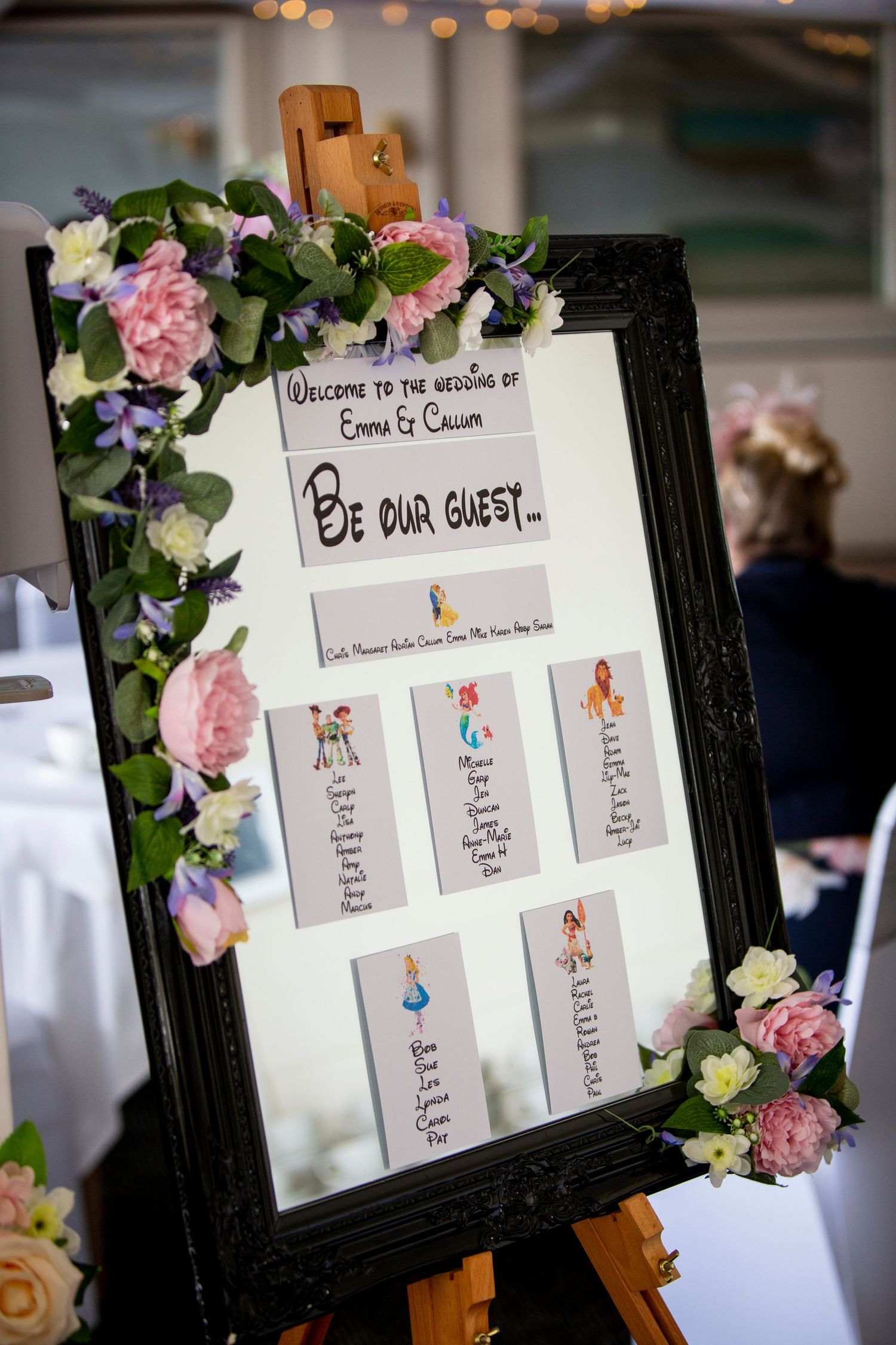 Wedding seating chart display with floral decorations in a black frame.