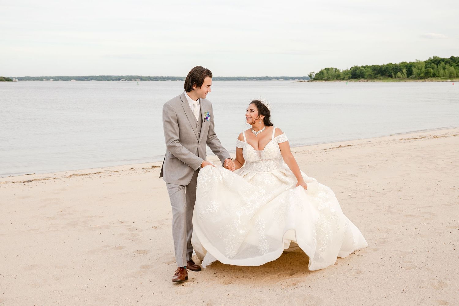 groom helping a bride carryng her dress while they walk on the beach sand in Greentree Country Club westchester NY