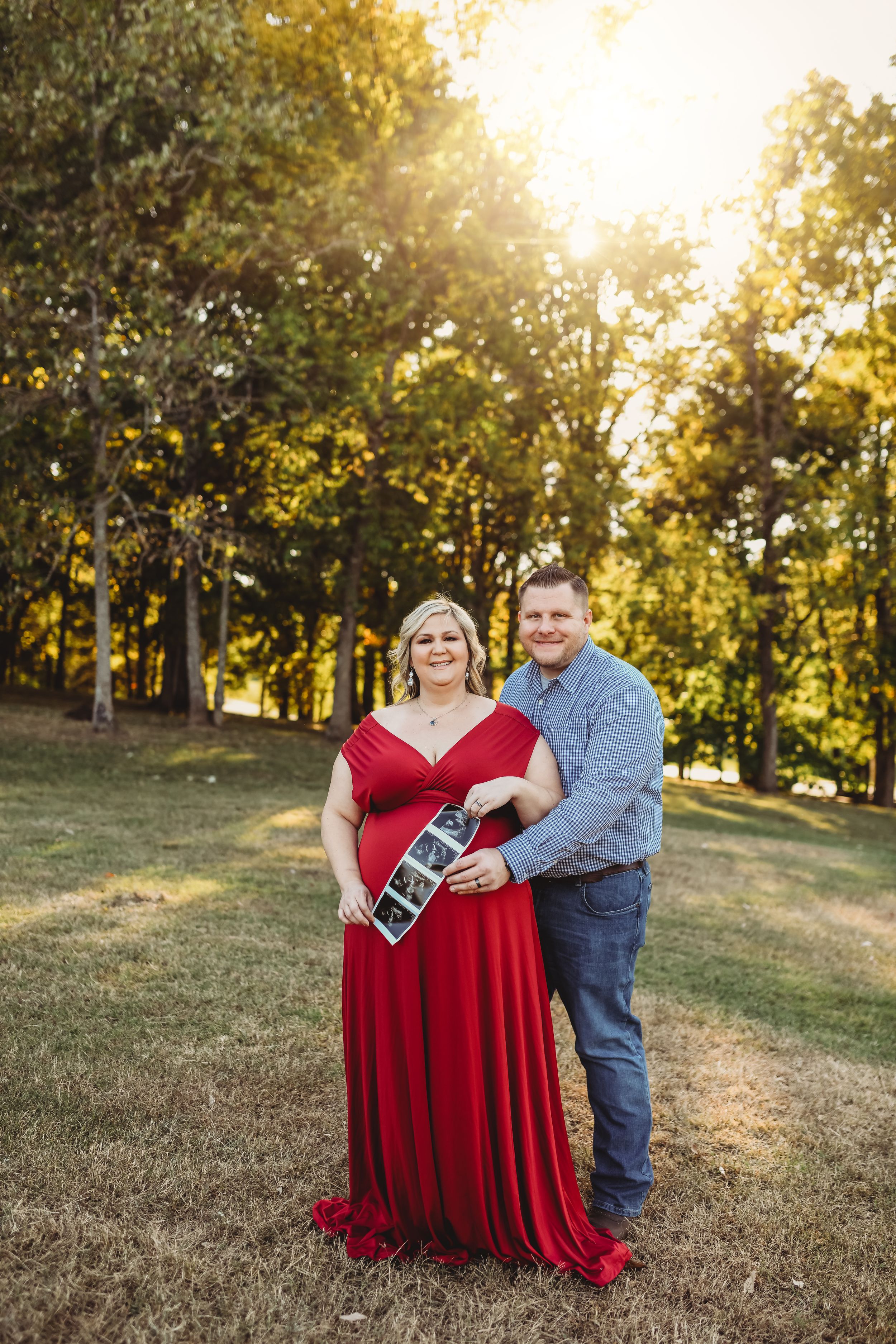 A couple poses for a maternity photo in a park at sunset holding ultrasound images.