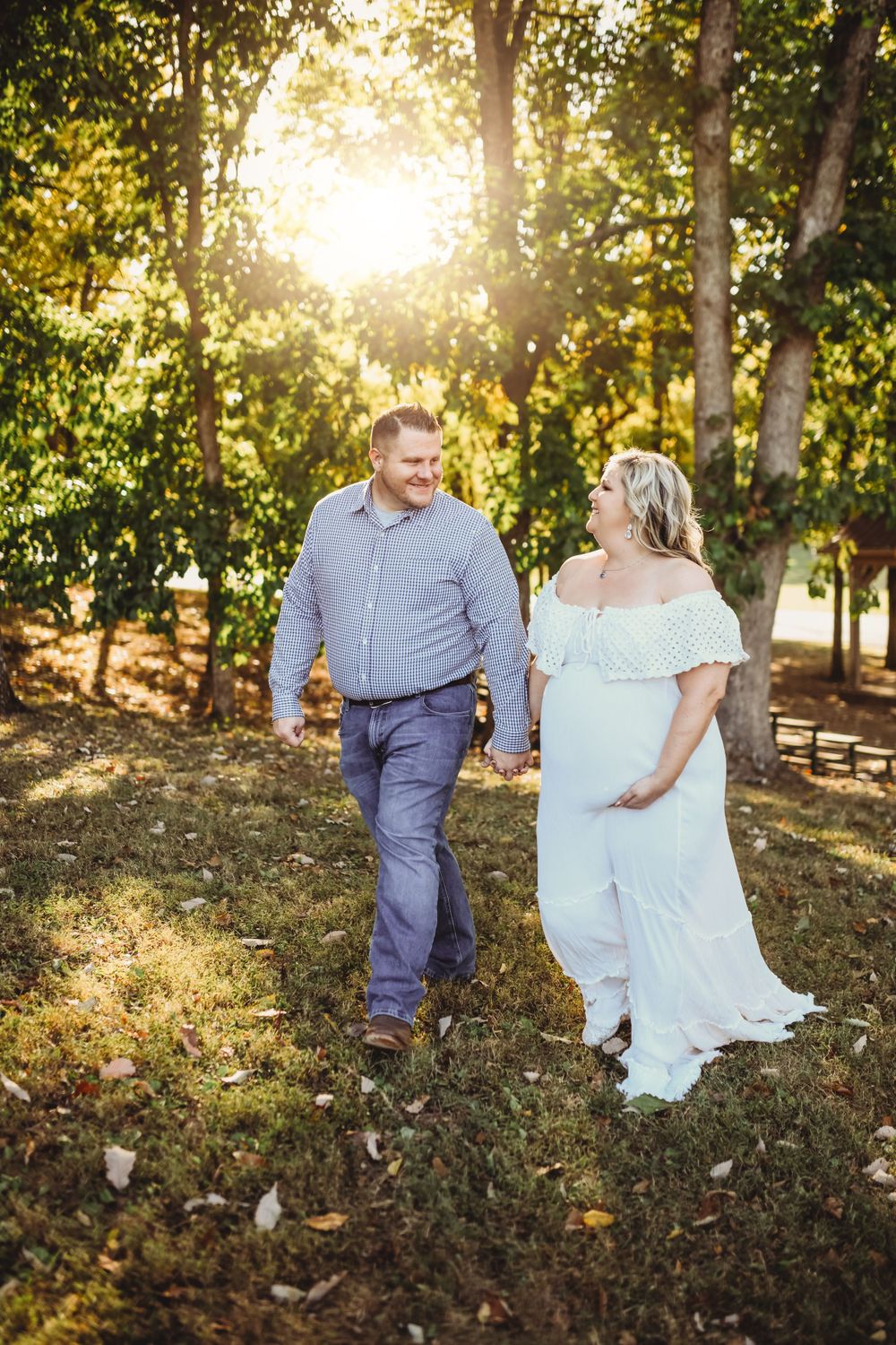 A couple walks hand in hand through a sunlit wooded area in formal attire.