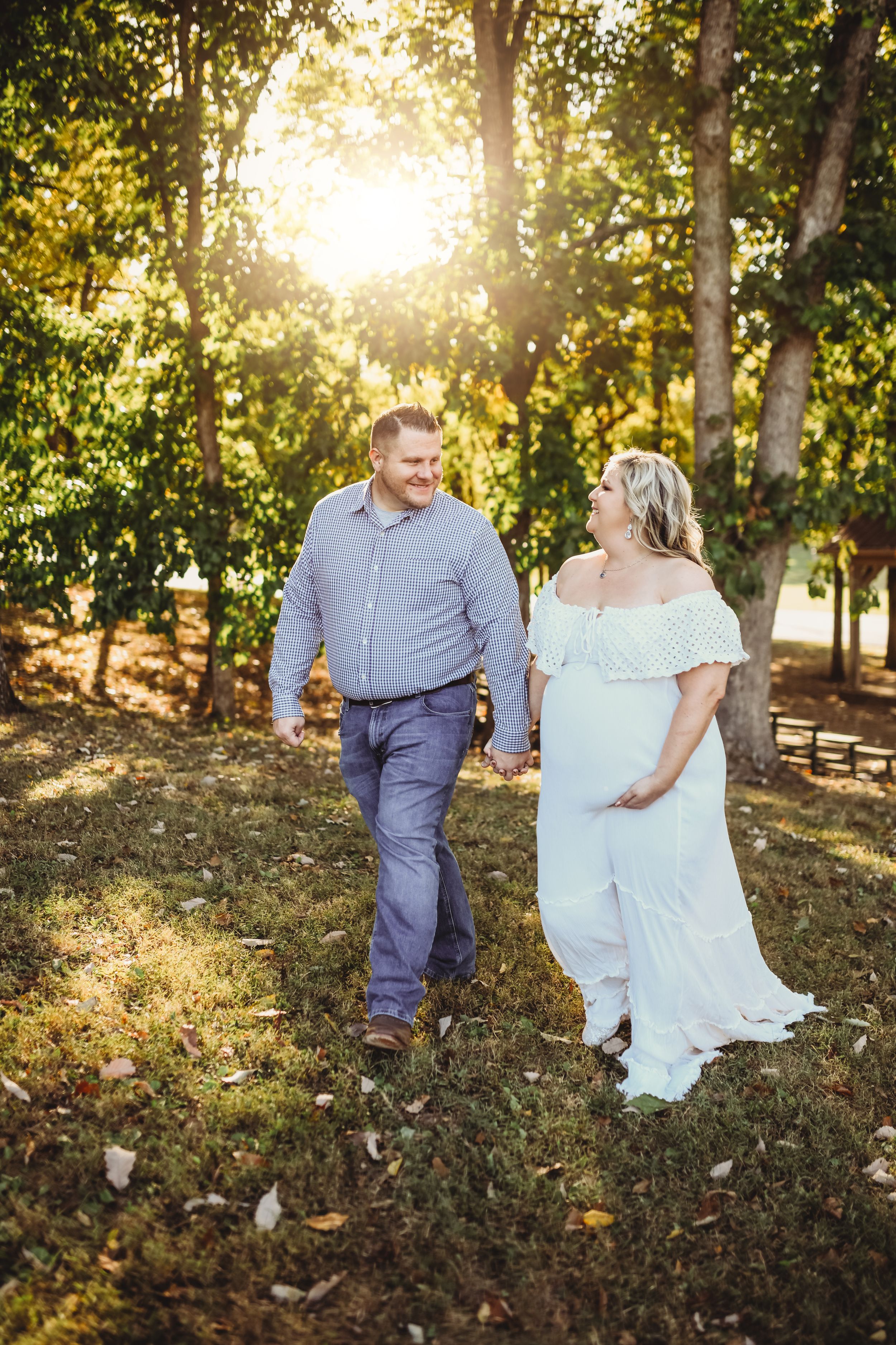 A couple walks hand in hand through a sunlit wooded area in formal attire.