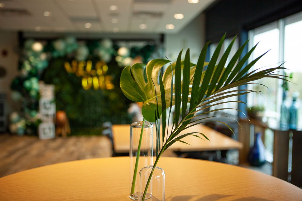 Tropical palm leaf in a glass vase on wooden table with green balloon decor in background.