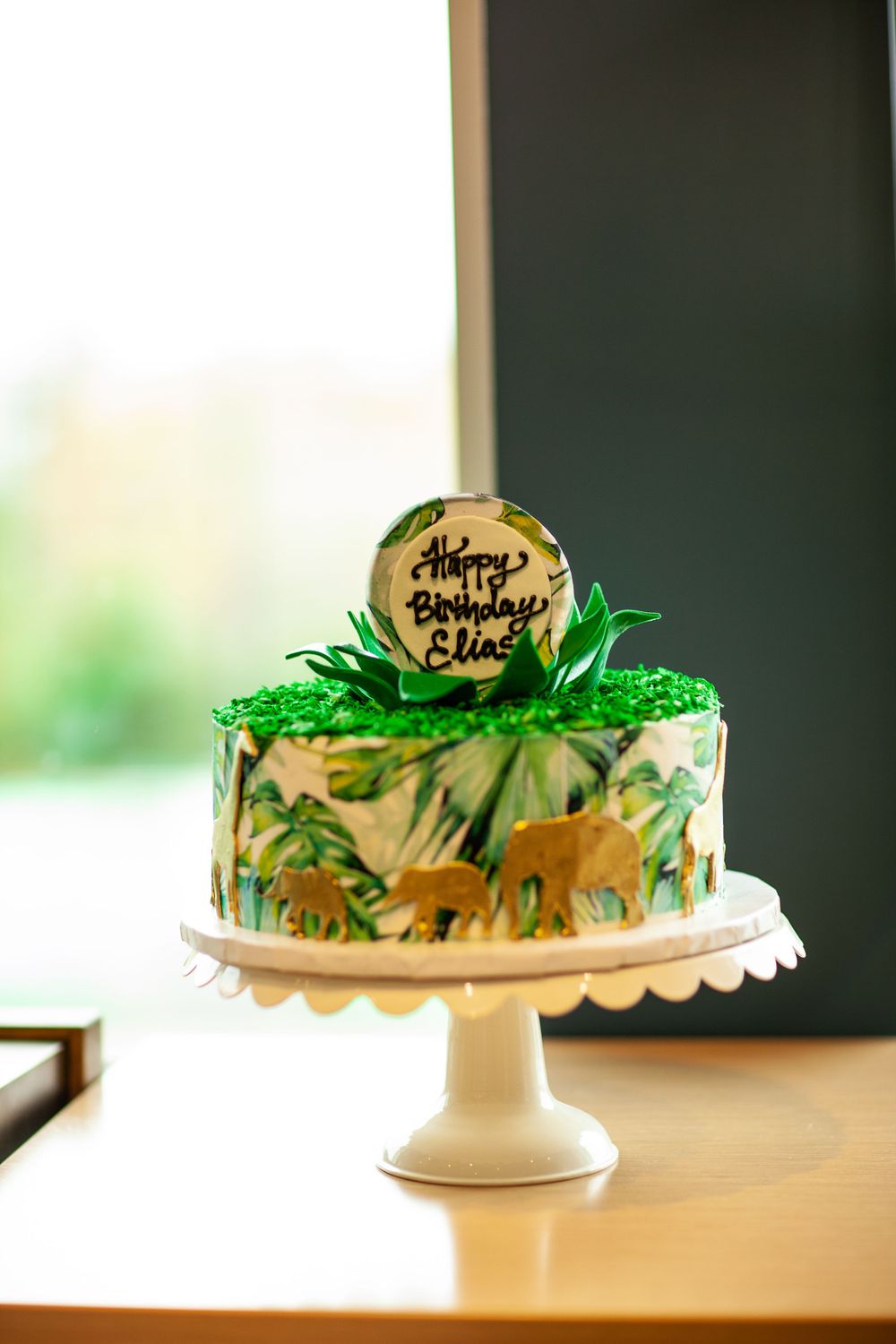 Safari-themed birthday cake decorated with green leaves and golden dinosaur silhouettes on white cake stand.