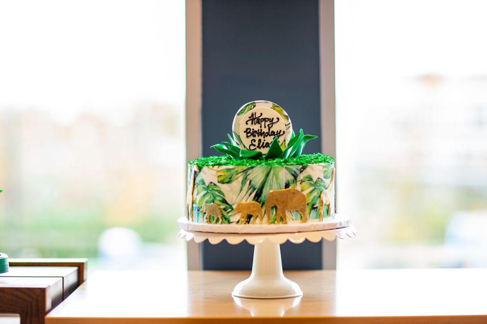 White birthday cake with jungle leaf decorations and gold birthday message on a pedestal stand.
