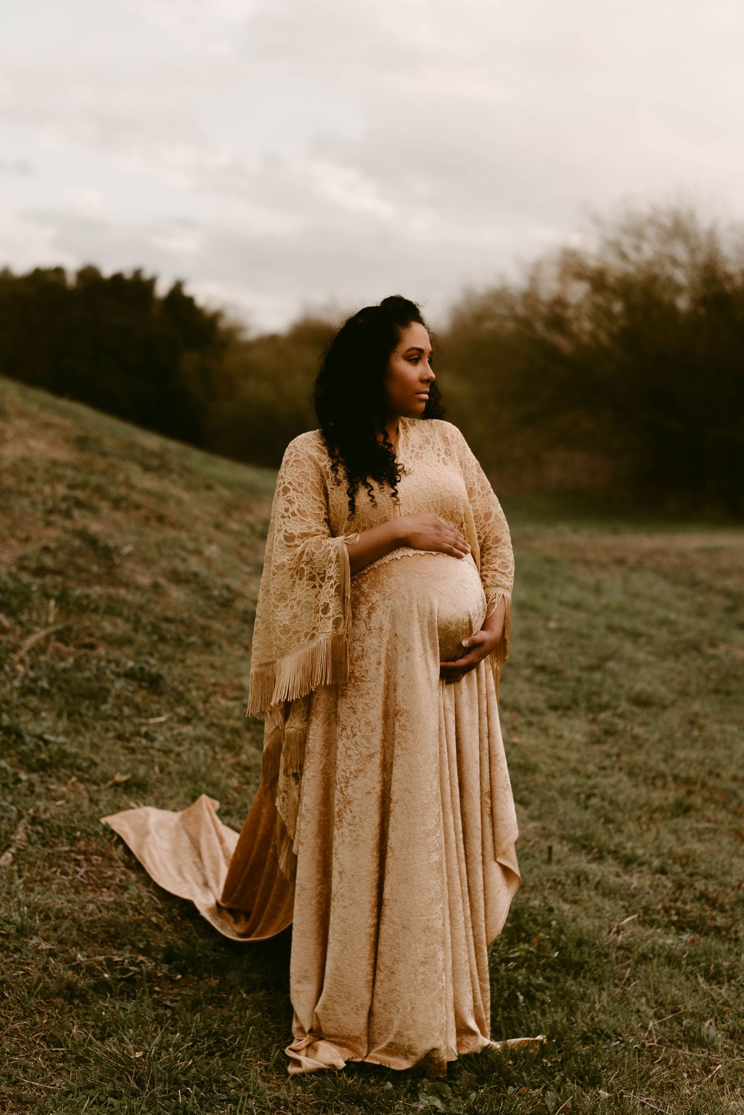 Warm-toned maternity portrait featuring lace dress with dramatic sleeves on hillside.