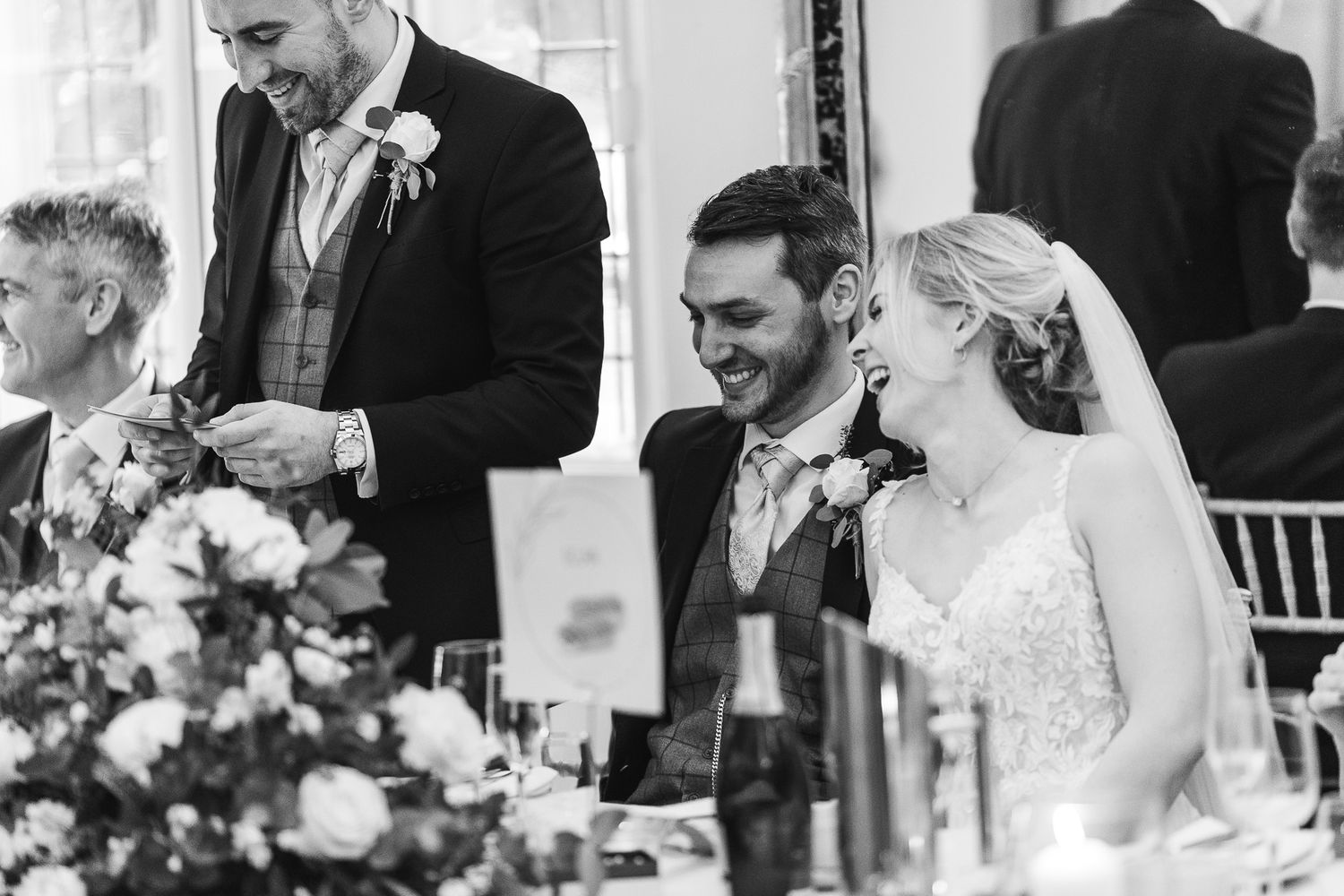 Black and white wedding reception image shows guests laughing at head table.