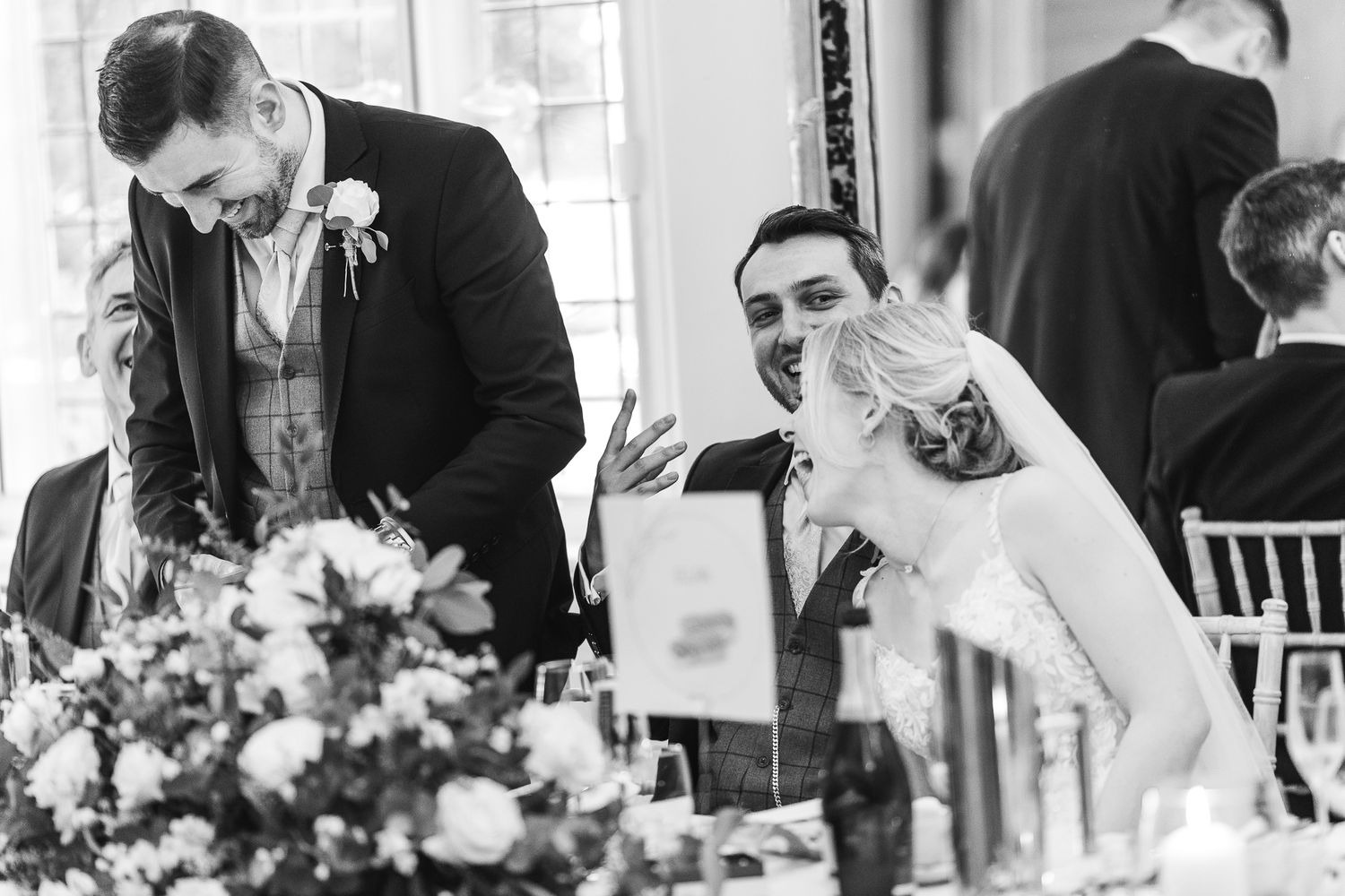 Candid black and white photo captures laughter and joy during wedding reception speeches.