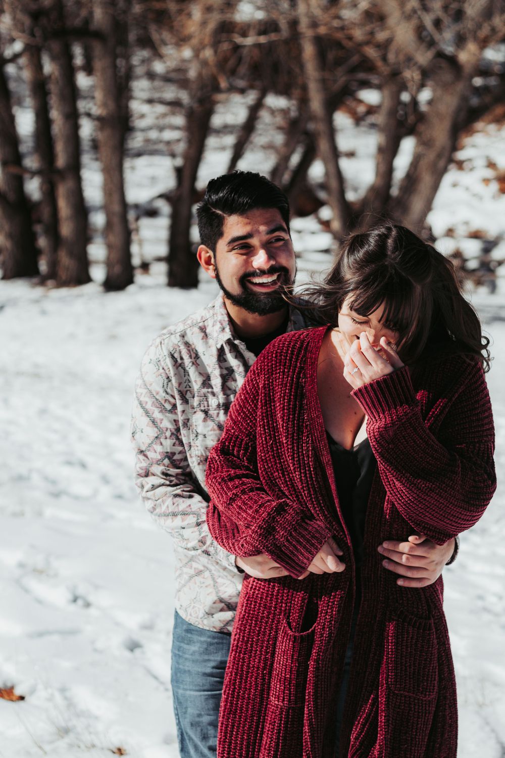 Playful winter portrait of couple in snow with red sweater and joyful expressions.