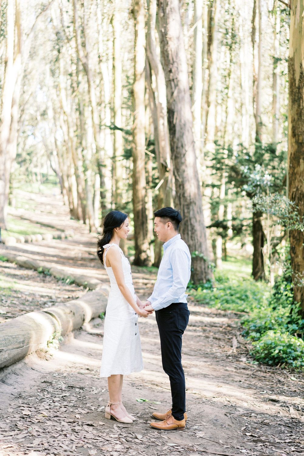 A couple holding hands on a dirt trail surrounded by towering trees in a serene forest setting.