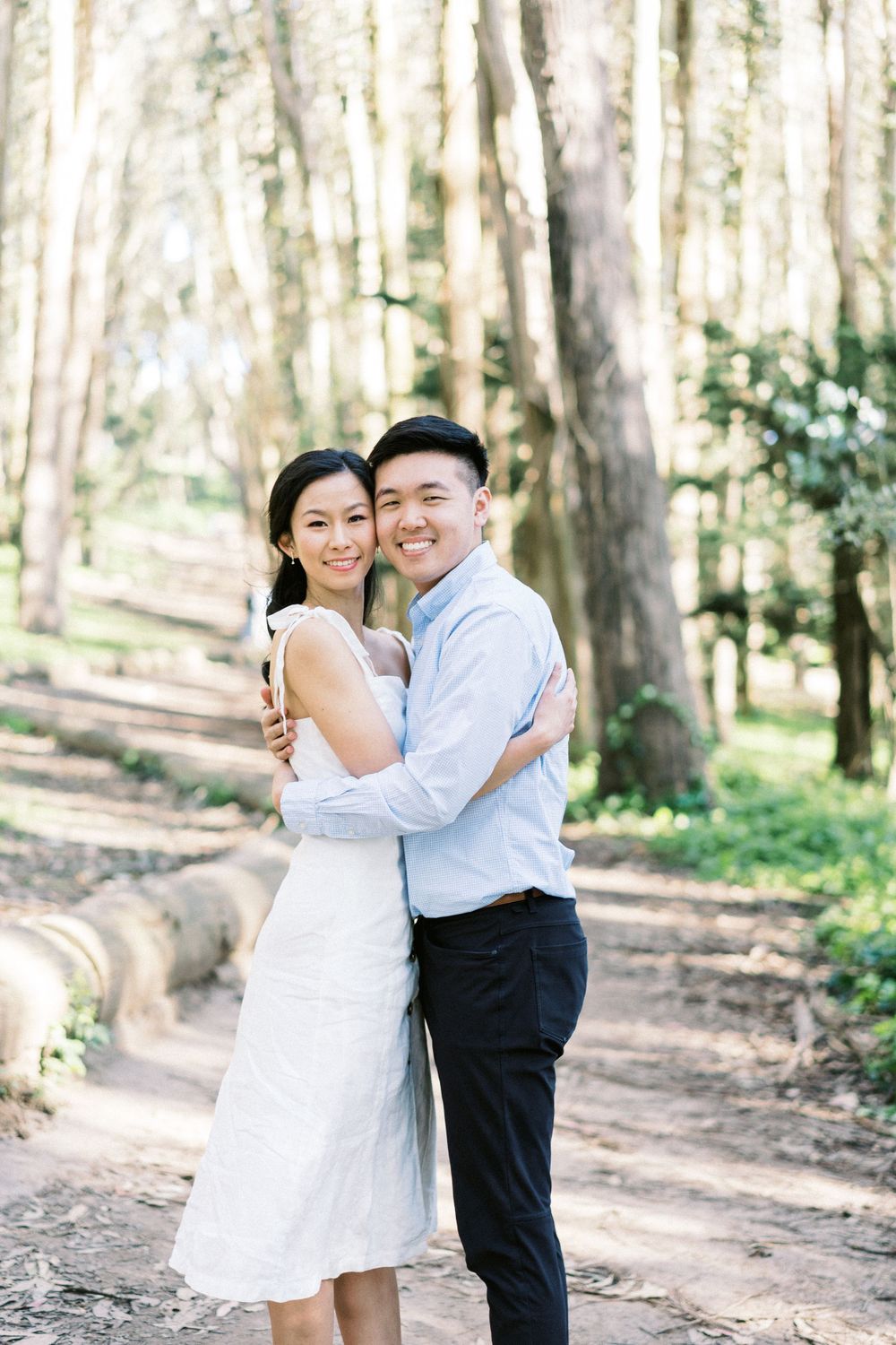 A happy couple embracing on a wooded path with sunlight filtering through the trees.