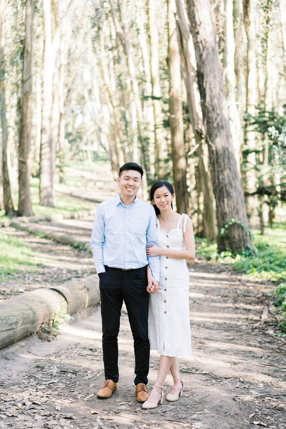 A couple standing together on a forest path with sunlight streaming through the trees.