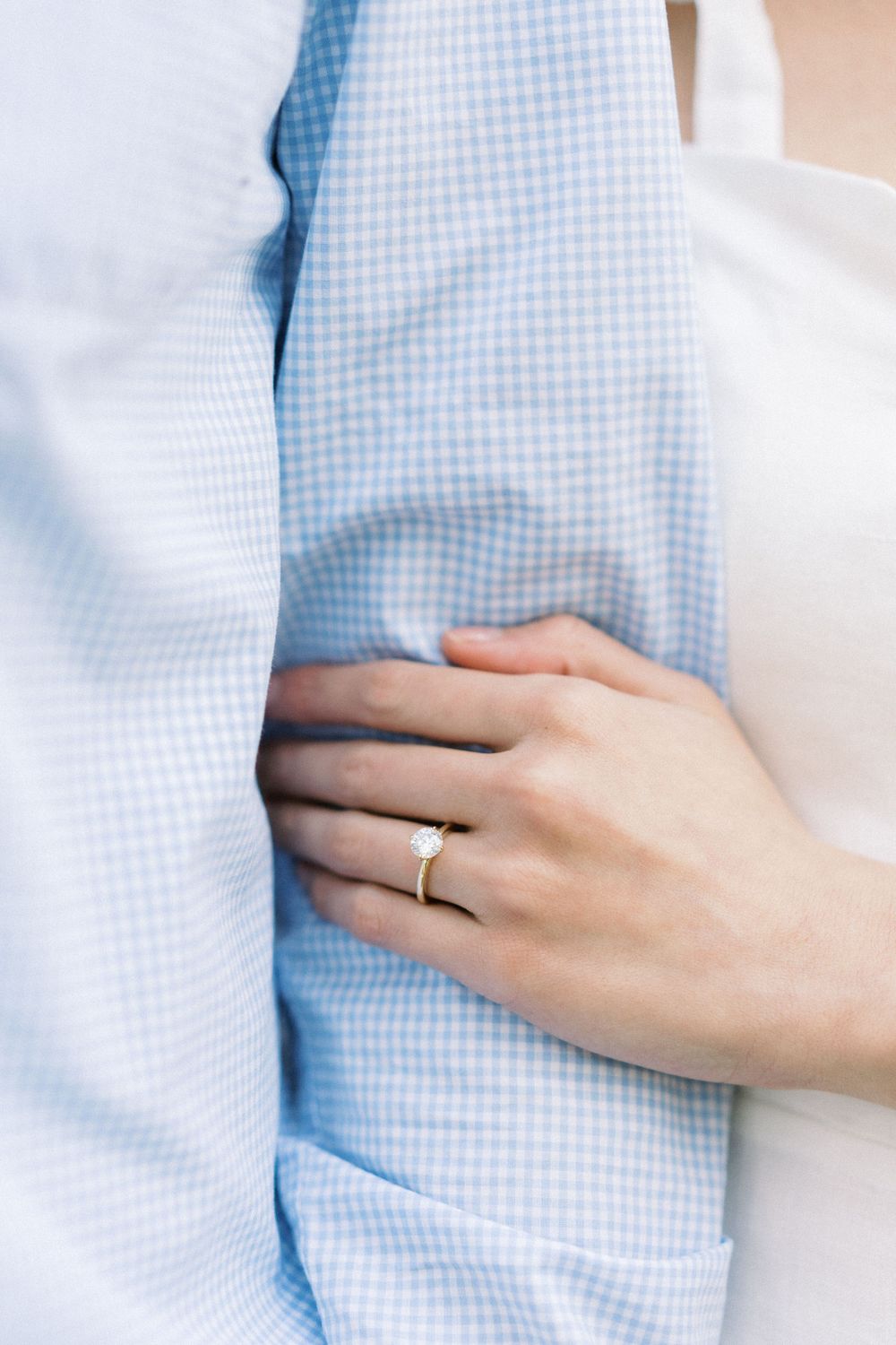 A close-up of hands showing an engagement ring against a light blue gingham shirt.