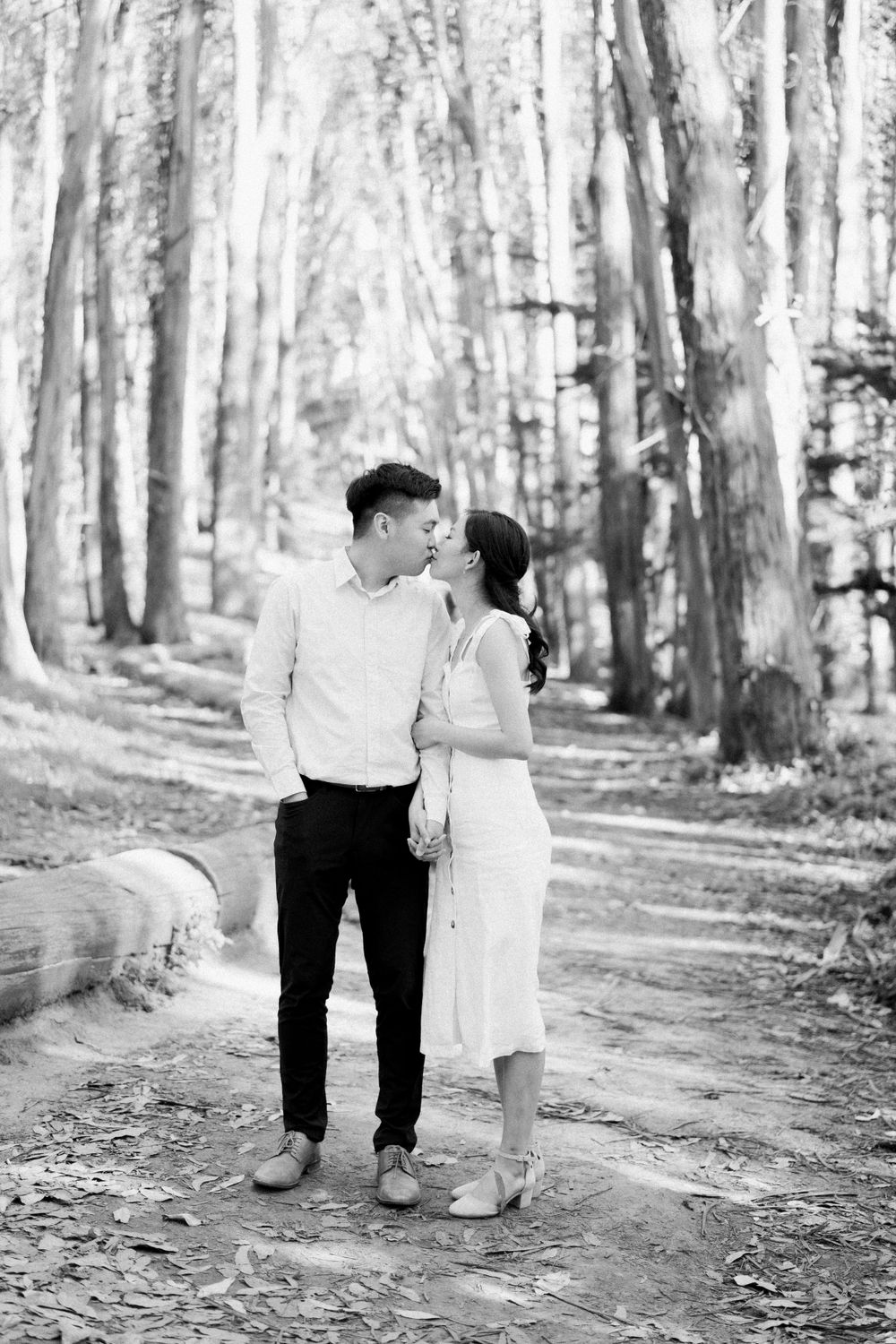 A romantic black and white portrait of a couple embracing on a wooded path surrounded by tall trees.