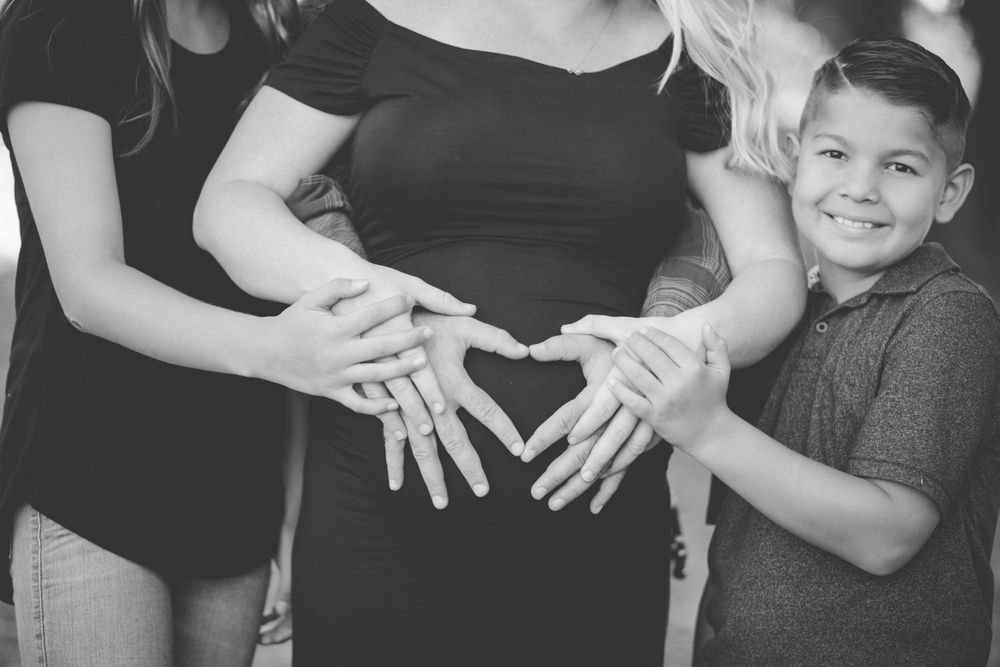 Hands form heart shape on pregnant belly with child joining in black and white photo.
