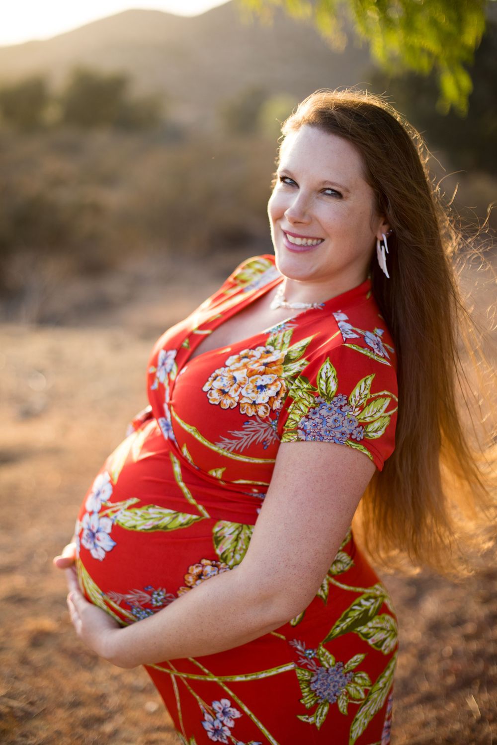 Expectant mother in red floral dress poses outdoors during golden hour sunset.