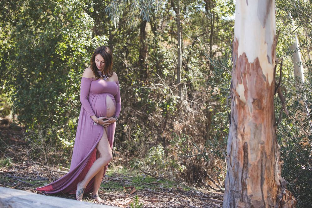 Pregnant woman in purple dress poses near eucalyptus tree in natural setting.