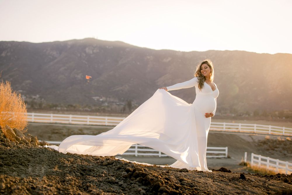 A flowing white wedding dress billows in the breeze at sunset with mountain and white fence backdrop.
