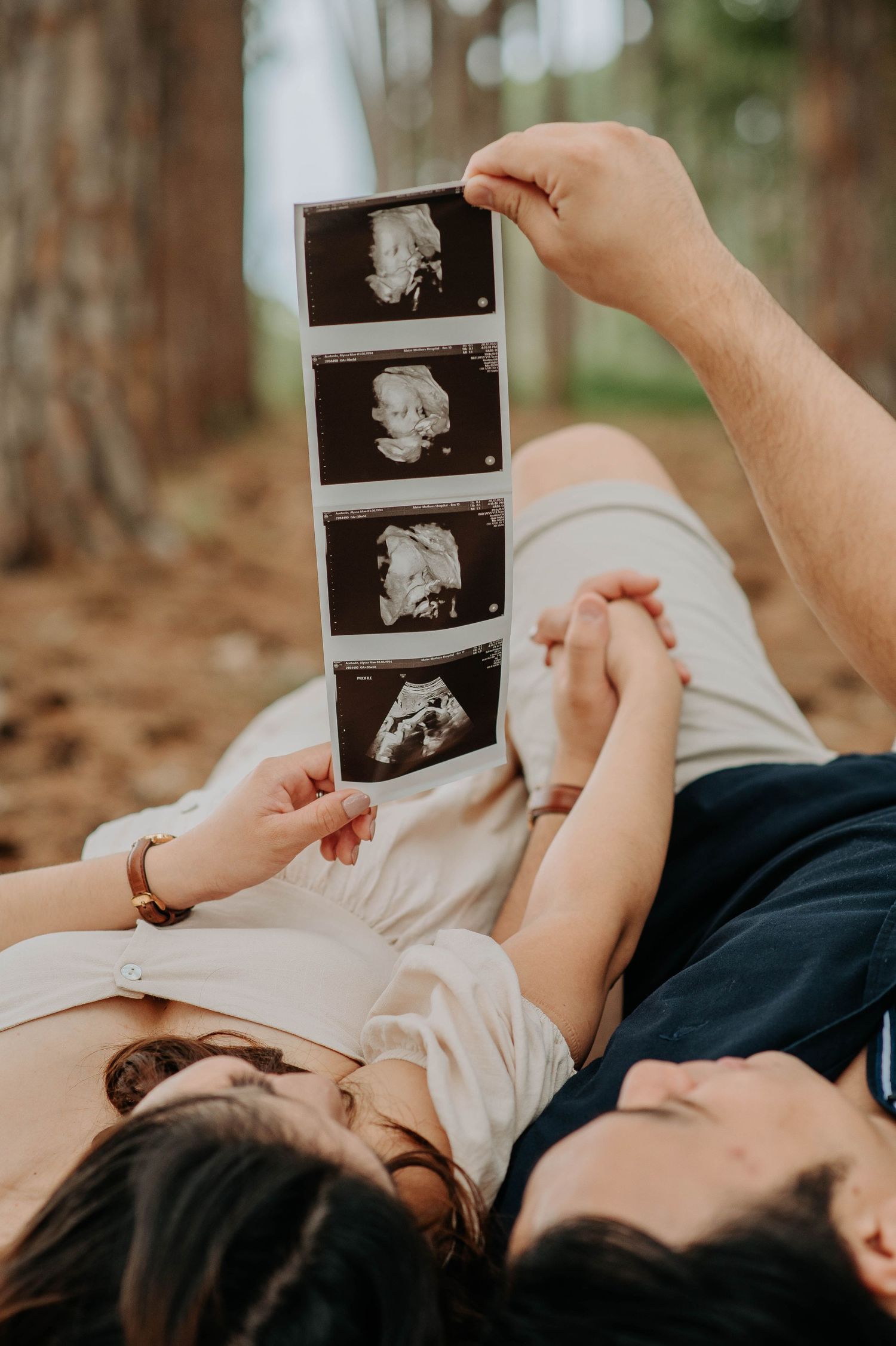 A couple lying down and holding ultrasound images in a forest setting.