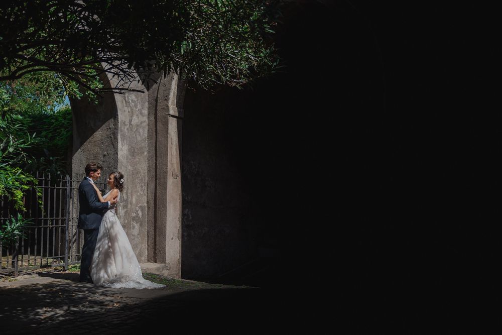 Romantic wedding portrait in stone archway with dramatic lighting and green foliage.