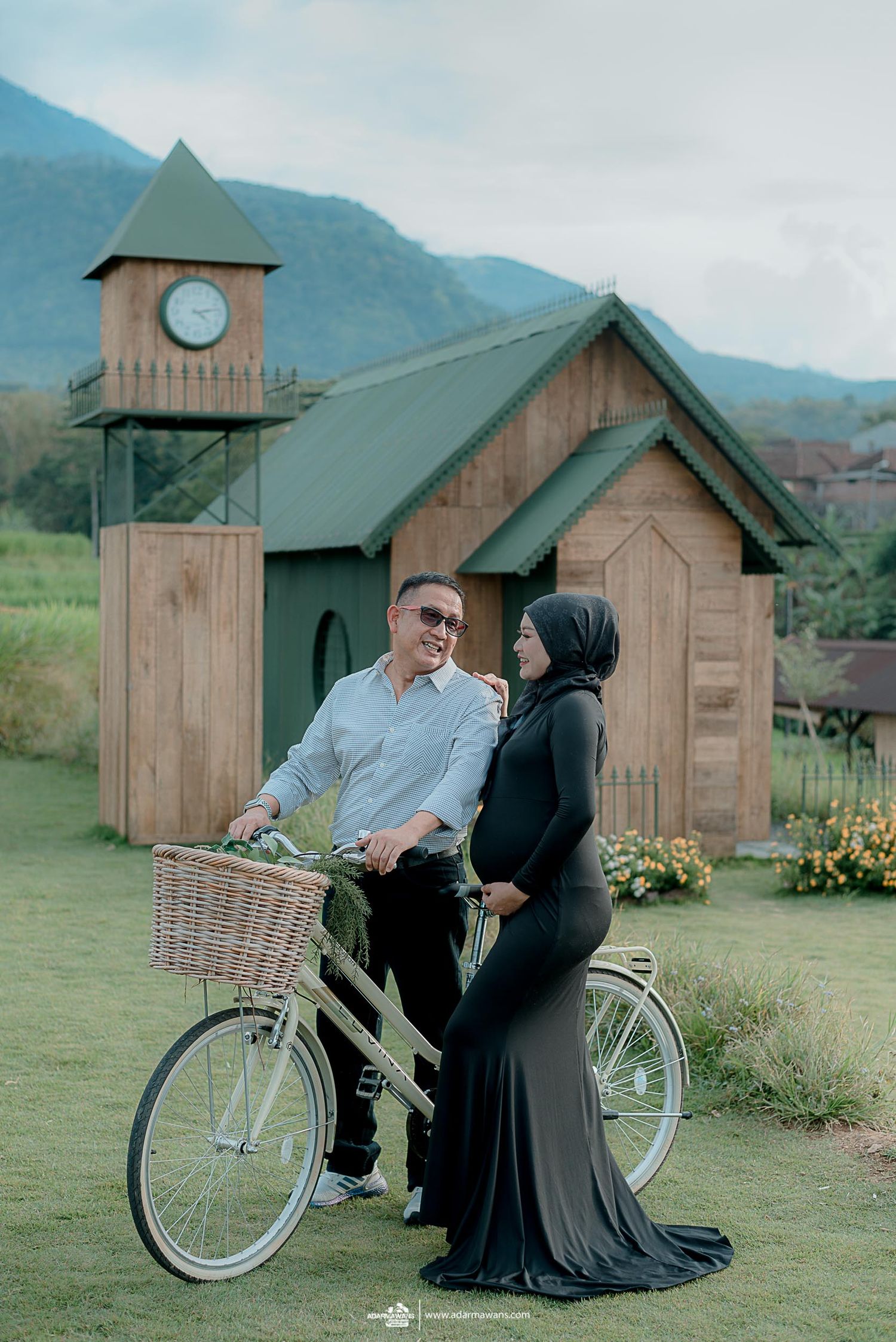 Couple posing with vintage bicycle in front of rustic wooden chapel with clock tower in mountain setting.