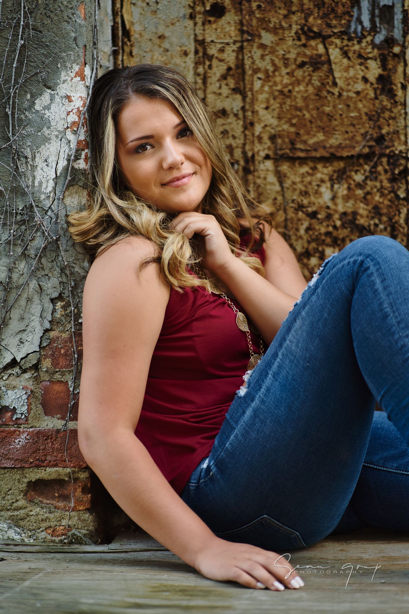 Casual portrait in burgundy top and jeans against rustic wall.