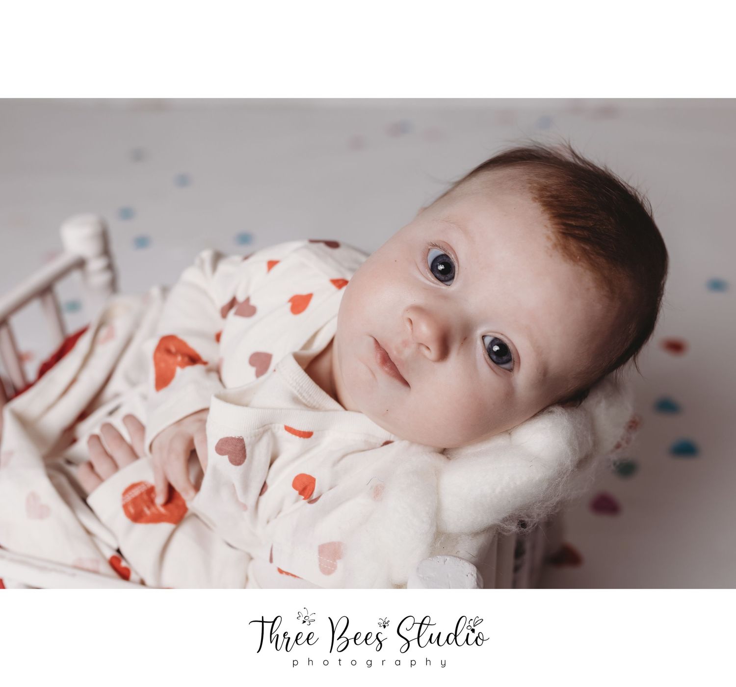 Baby wearing heart print outfit lying in vintage crib with soft lighting.