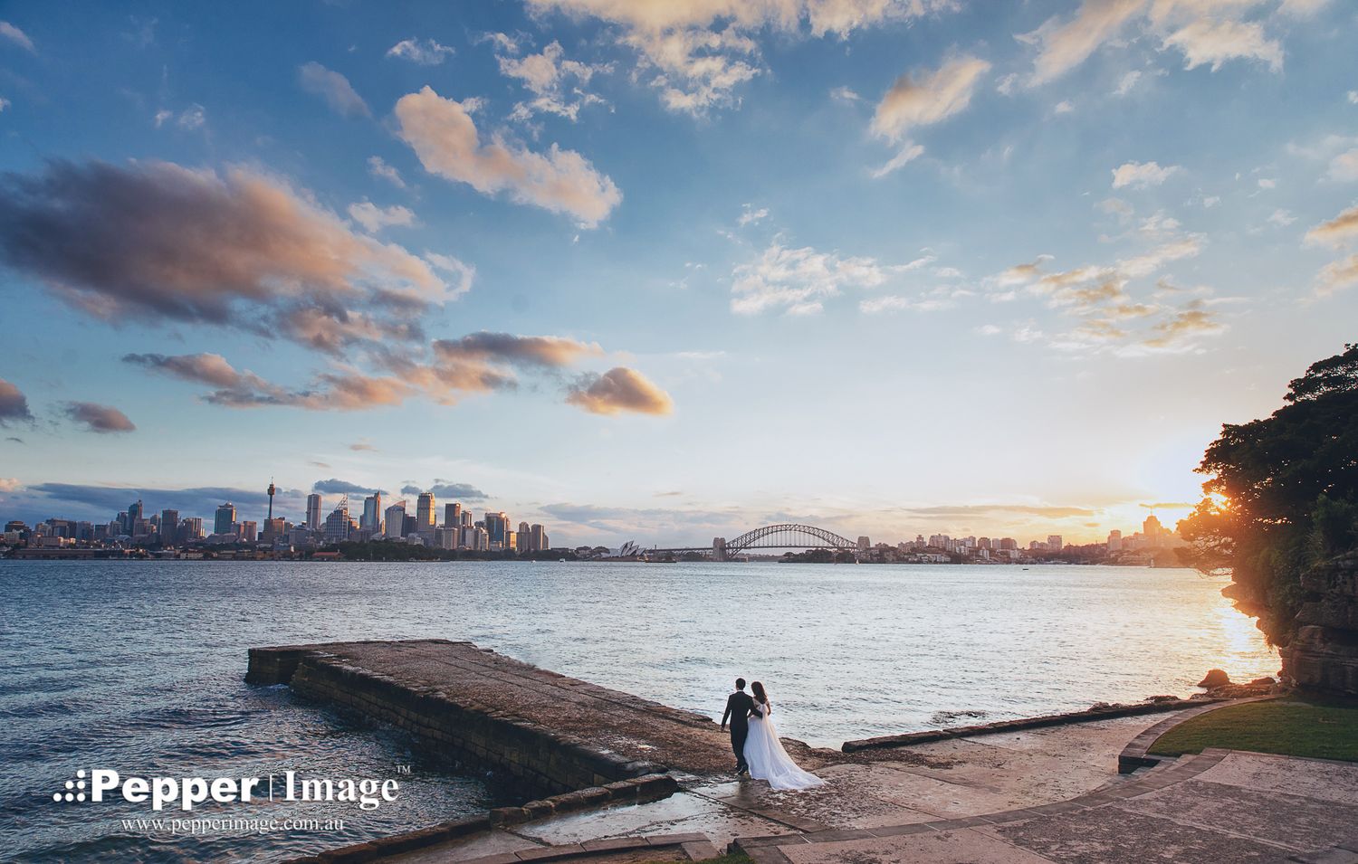 Bride and groom share a romantic moment by the waterfront with Sydney Harbour Bridge at sunset.